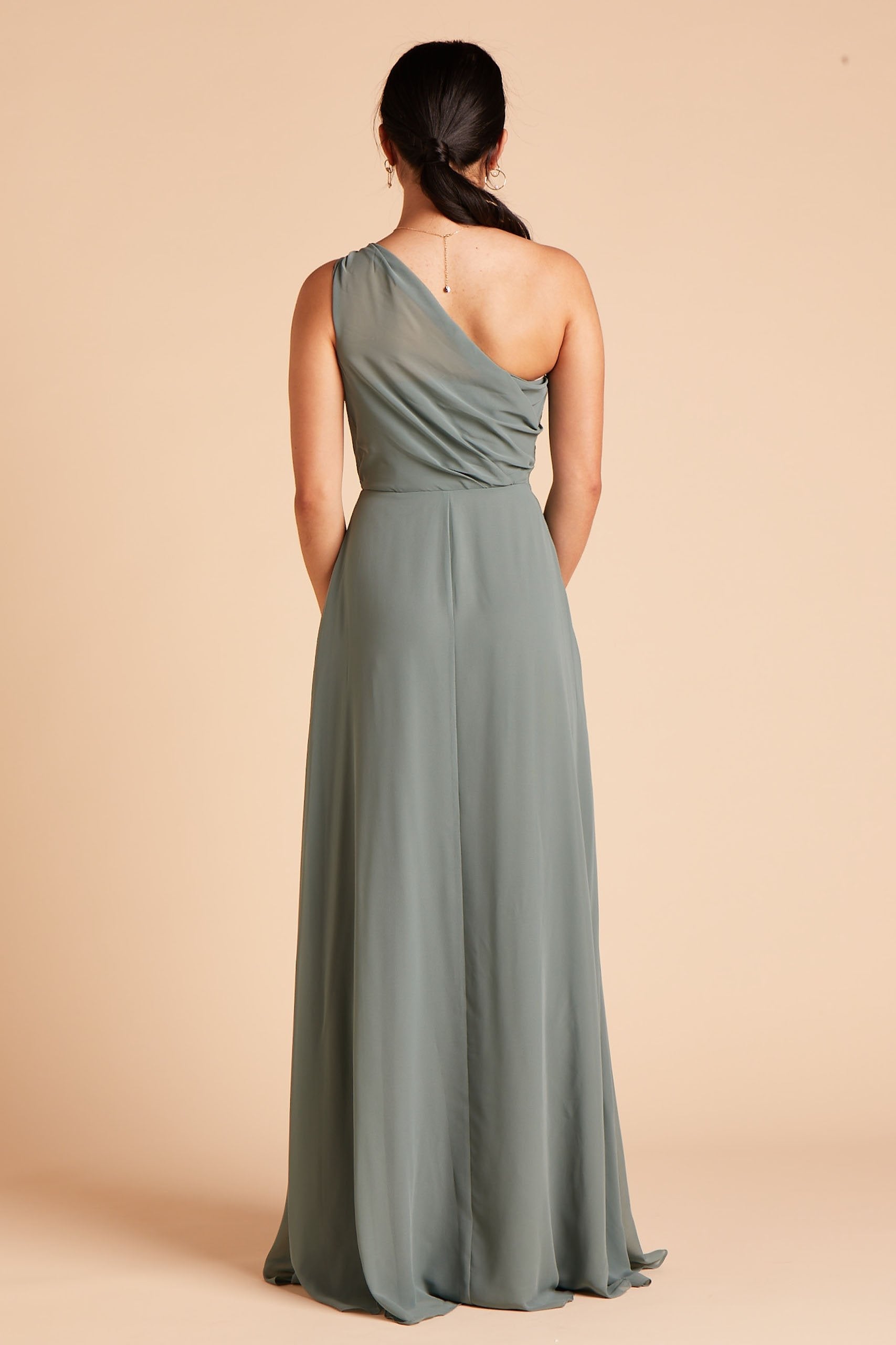 Back view of the Kira Dress in sea glass chiffon shows a model wearing sheer, softly pleating chiffon gathered at the bodice shoulder and draping across the back to the side seam as the dress flows to the floor. 