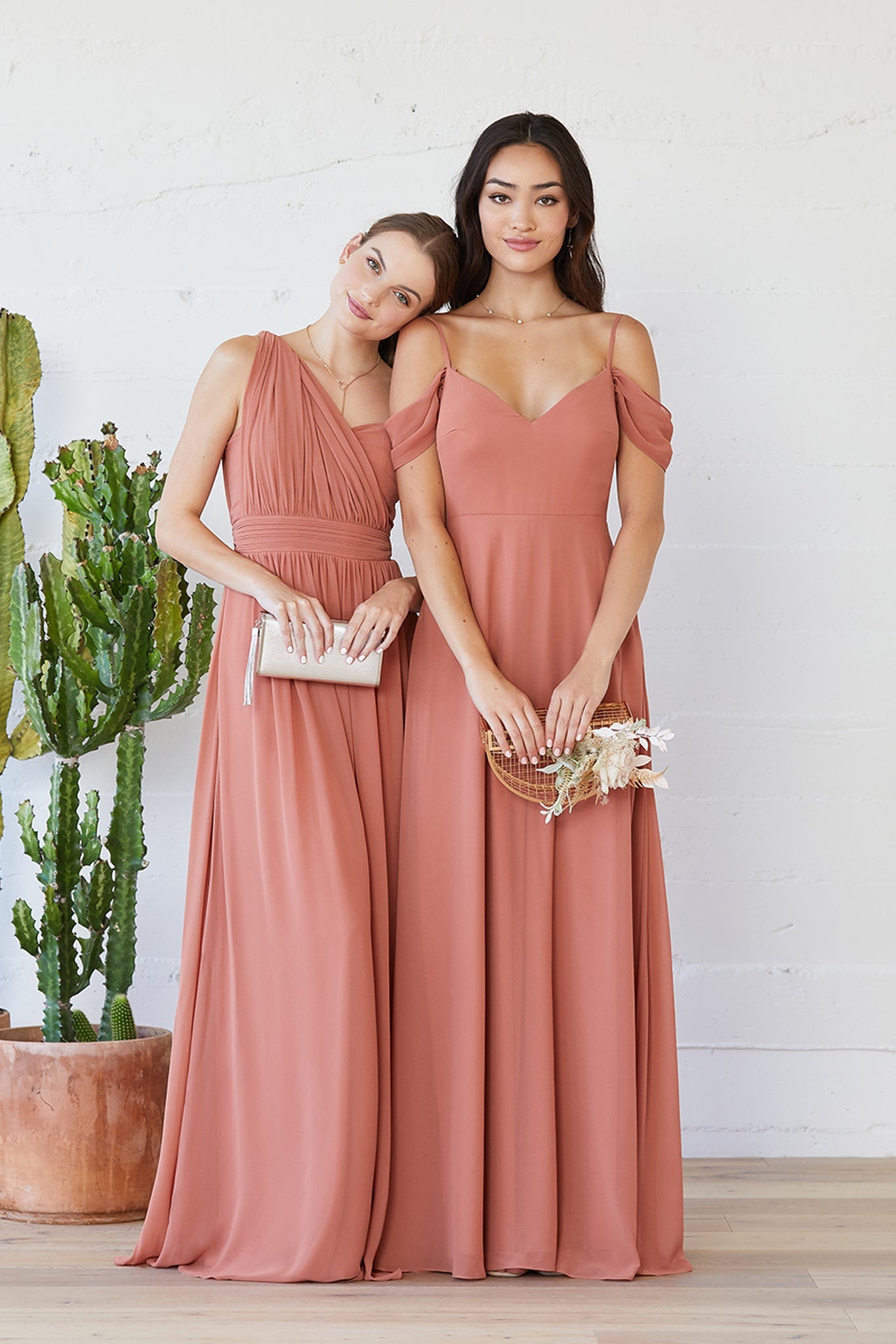 Two models stand together wearing terracotta chiffon bridesmaid dresses. On the left, a slender model with light skin wearing a Grace Convertible Bridesmaid Dress rests her head on the shoulder of a model wearing a Devin Convertible Bridesmaid Dress.