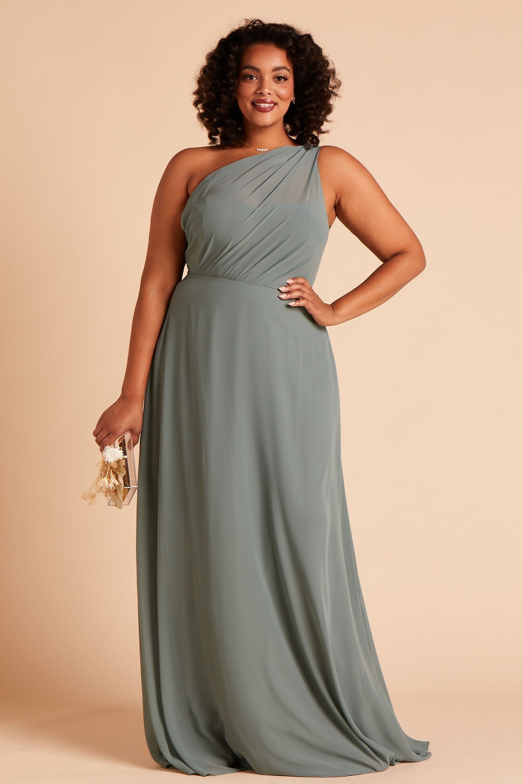 Front view of the Kira Dress Curve in sea glass chiffon without the optional slit shows a full-figured model with a medium skin tone wearing an asymmetrical one-shoulder, full-length dress. Soft pleating gathers at the left shoulder of the bodice with a smooth fit at the waist as the dress with a slight A-line silhouette flows to the floor.