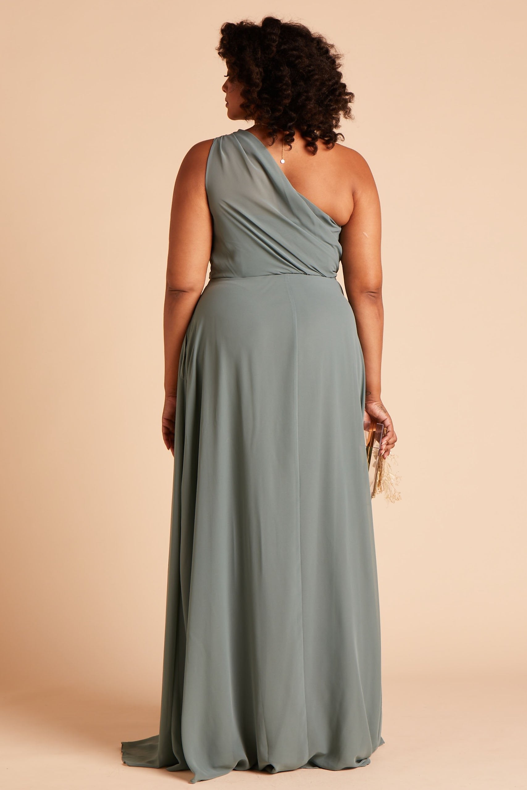 Back view of the Kira Dress Curve in sea glass chiffon shows a full-figured model with a medium skin tone. The back of the dress has sheer chiffon fabric which creates a pleated Grecian one-shoulder neckline and bodice. 